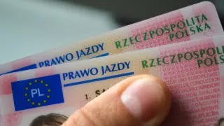 Dwa polskie prawa jazdy, jedno na drugim. Osoba trzyma je w dłoni, gotowa do jazdy w okularach bez wpisu.
