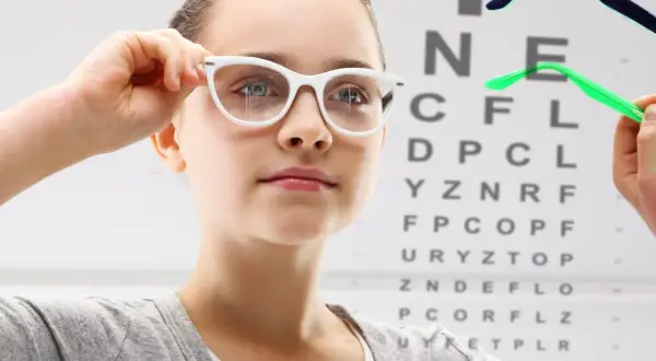 Dziewczynka przymierza okulary, sprawdzając co na poprawę wzroku. W tle tablica Snellena.