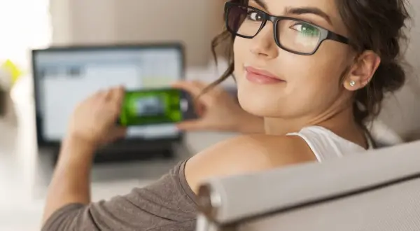 Kobieta w okularach z lekko zmęczonymi oczami trzyma telefon przed laptopem.