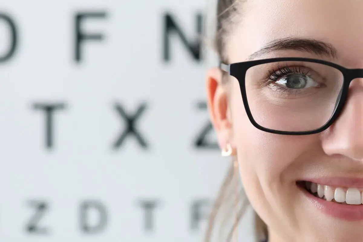 Uśmiechnięta kobieta w okularach przed tablicą Snellena. Kto to jest optometrysta? Specjalista od wzroku, który dba o Twoje oczy.