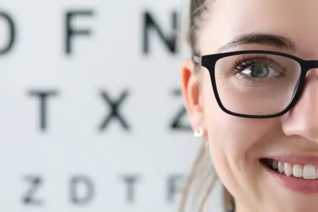 Uśmiechnięta kobieta w okularach przed tablicą Snellena. Kto to jest optometrysta? Specjalista od wzroku, który dba o Twoje oczy.